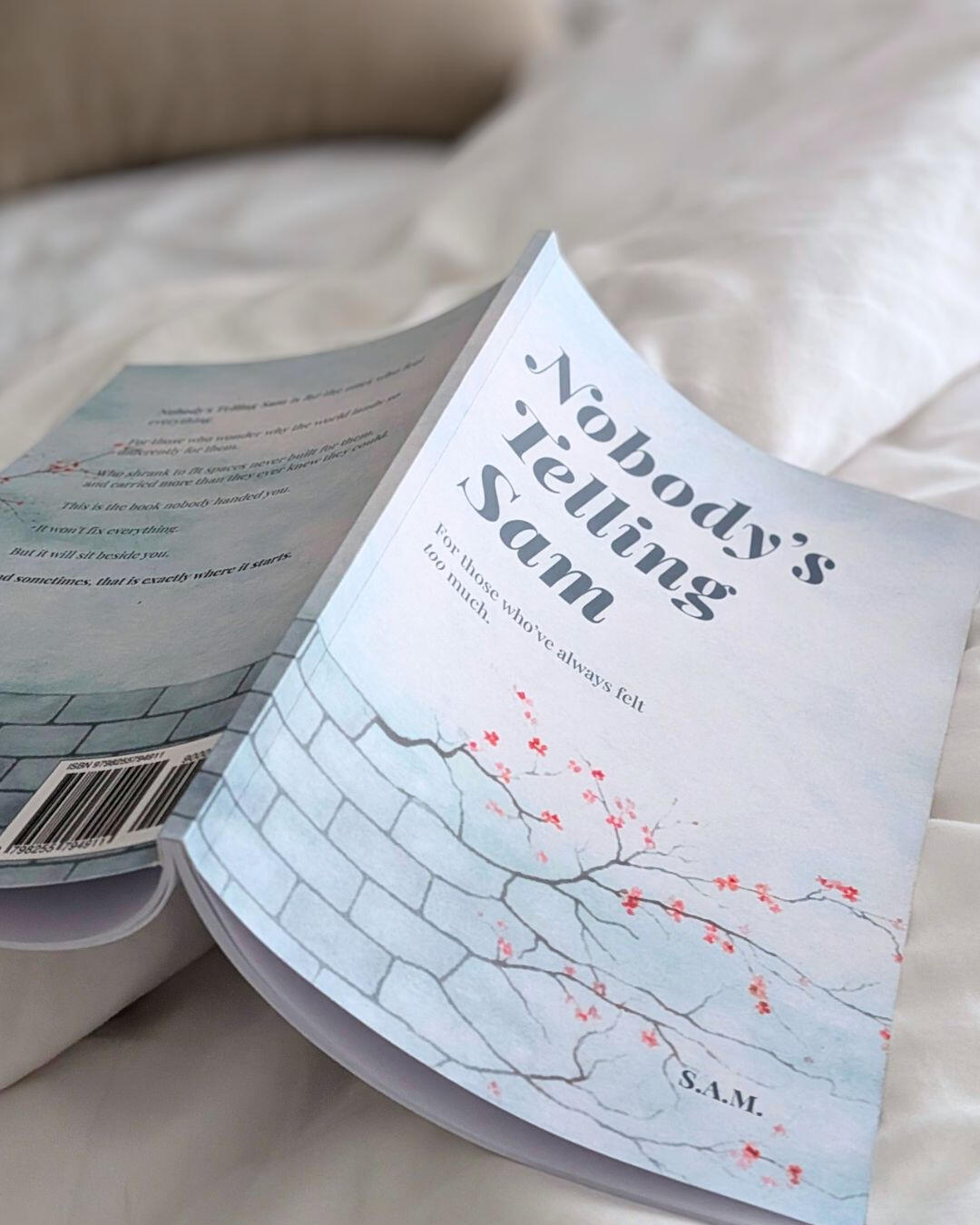 A soft-focus photograph of the book 'Nobody’s Telling Sam' lying open on rumpled white cotton bedsheets, capturing a sense of quiet, intimate reflection.