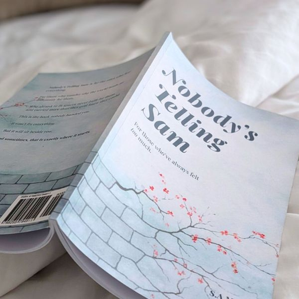 A place to rest when the world feels like too much. A soft-focus photograph of the book 'Nobody’s Telling Sam' lying open on rumpled white cotton bedsheets, capturing a sense of quiet, intimate reflection.