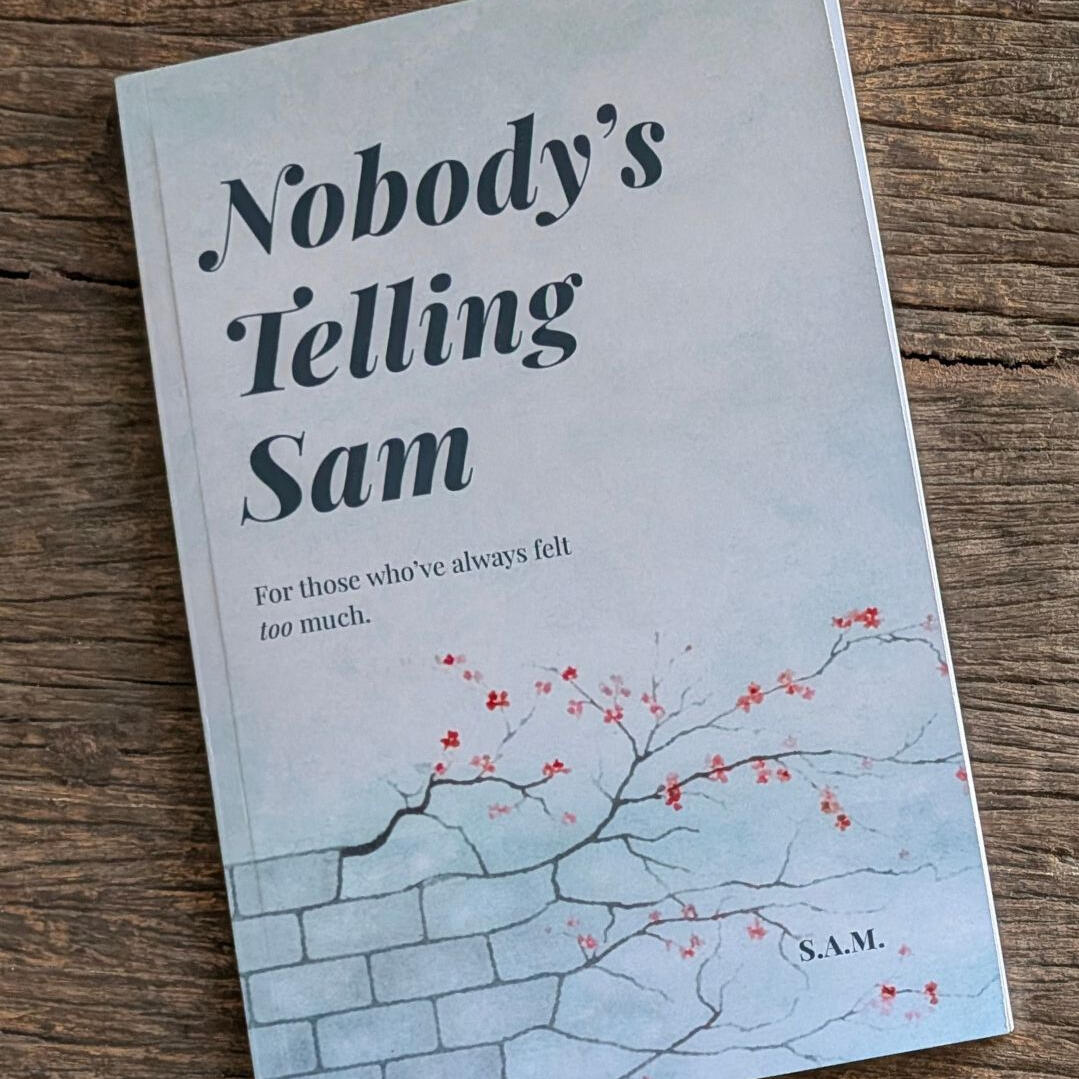 The front cover of the book 'Nobody’s Telling Sam' by S.A.M., resting on a textured dark wood surface, featuring a soft blue background with delicate red floral branches.